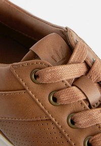 Bruna lädersneakers med vävda snören, perforerad sidodekoration, metallöglor och präglad logotyp på tungan. Slät textur.