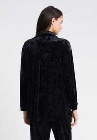 Camisa de terciopelo negro, de mangas largas, corte relajado, detalle de pliegue en la parte posterior, textura suave, brillo sutil. Combinada con pantalones de terciopelo a juego.