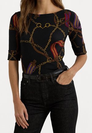 Kobieta ubrana w czarną dopasowaną bluzkę ze złotym łańcuchem, skórzanym paskiem oraz paskami w czerwono-fioletowe paski, w połączeniu z czarnymi dżinsami i paskiem.