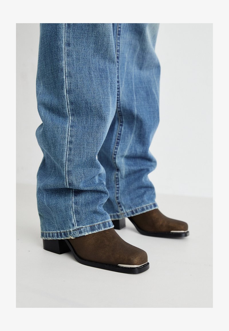 Botas de tornozeleiro de couro castanho com biqueira quadrada, saltos pretos e detalhes em prata, combinadas com jeans denim azul claro de corte relaxado.