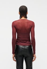 Άτομο με πλεξούδες, φορώντας διαφανές μακρυμάνικο μπορντό τοπ και μαύρο δερμάτινο παντελόνι, στέκεται με την πλάτη γυρισμένη σε απλό φόντο.