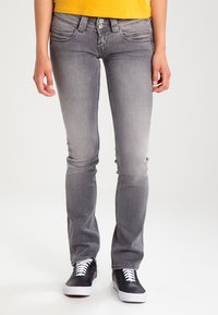 Vaqueros de mezclilla gris con un corte recto, desvanecimiento sutil, diseño de cinco bolsillos y cierre de dos botones, usados con zapatillas negras.