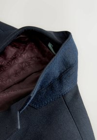 Colletto di blazer blu navy alzato, che mostra la fodera interna con motivo viola scuro e il tessuto strutturato sul colletto e sulla spalla.