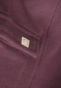 Lilla strikket stof med en blød tekstur, prydet med et lille cremefarvet mærke med et bjørnedesign syet på en lomme.