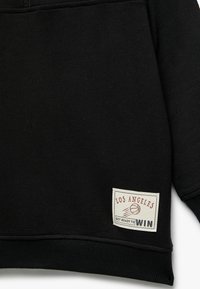 Zwarte sweatshirt met een geribbelde onderrand en manchetten, met een rechthoekig wit label op de linkeronderkant, waarop "LOS ANGELES" en "GET READY TO WIN" staat.