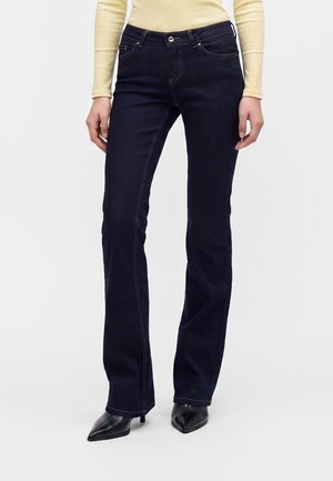 Person iført mørkeblå straight-leg jeans, sorte spidse sko og en lysegul bluse med lange ærmer mod en hvid baggrund.