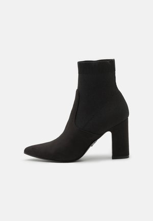 Schwarze Knöchelstiefel mit einem flexiblen Strick-Obermaterial, spitzer Zehenform und einem Blockabsatz, die eine glatte, samtige Textur aufweisen.