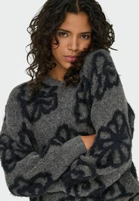 Frau mit lockigem, dunklem Haar, die einen grauen, flauschigen Pullover mit großen schwarzen Blumenmustern trägt und ihre Arme eng an die Brust hält.