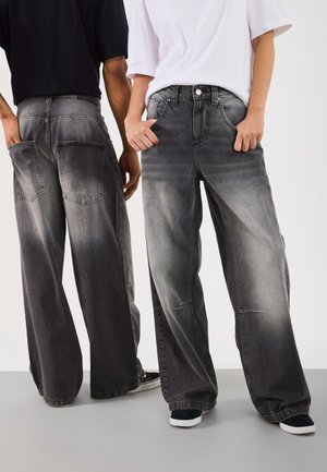 Zwei Personen tragen weit geschnittene ausgewaschene schwarze Jeans, eine mit dem Rücken zur Kamera in schwarzem Shirt, die andere zeigt nach vorne in weißem Shirt, Hände in den Taschen.