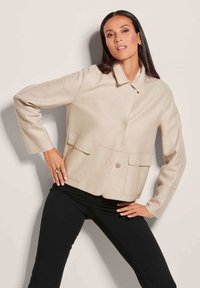 Beige cropped jack met een gestructureerde stof, voorzien van een klassieke kraag, een sluiting met knopen aan de voorkant en twee voorzakken. Draagt met zwarte broek.