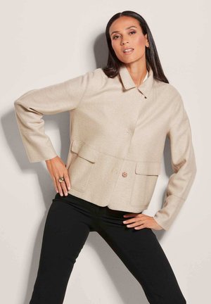 Beige cropped jack met een gestructureerde stof, voorzien van een klassieke kraag, een sluiting met knopen aan de voorkant en twee voorzakken. Draagt met zwarte broek.