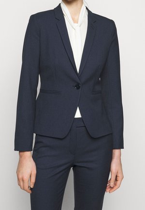 Vrouw draagt een nauwsluitend donkerblauw jasje met één knoop en bijpassende broek, gecombineerd met een witte blouse die bij de hals is gestrikt.