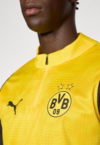 Κίτρινο αθλητικό μπουφάν με φερμουάρ στο γιακά, που φέρει μαύρο λογότυπο Puma και στρογγυλή πλακέτα BVB 09 με αστέρια, σε υφασμάτινη υφή.