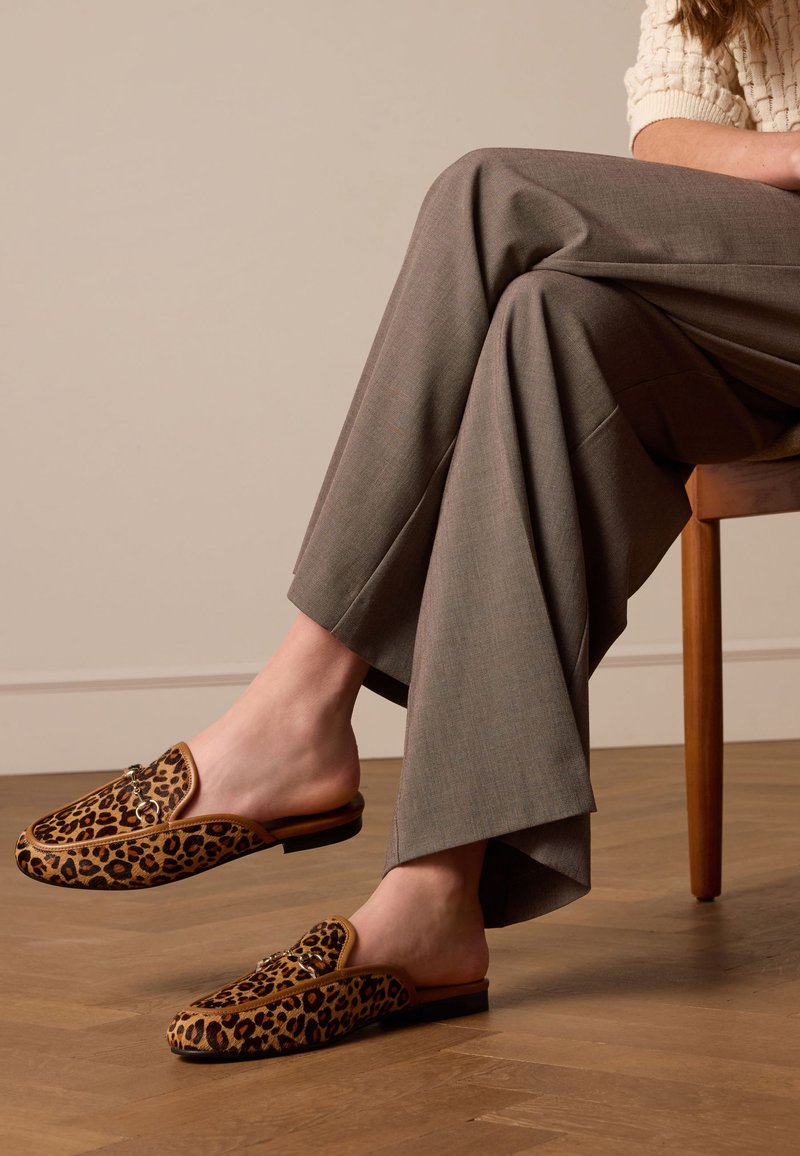 Leopard print mules with a soft, textured finish and a flat sole, paired with wide, gray trousers. Wood chair in the background.