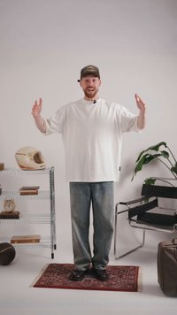 Mann in weißem oversized Hemd und Kappe steht mit offenen Armen in minimalistischem Raum mit Stuhl, Pflanze, Regal mit Helm, Büchern und Uhr.