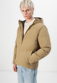 Tan puffer jacket with a hood, textured fabric, front zipper, and side pockets, worn over a cream sweater and paired with light blue jeans.