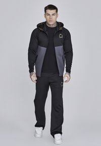 Černá a šedá mikina na zip s kapucí a logem, spárovaná s černými kalhotami s emblémem. Materiál vypadá hladce a těsně.