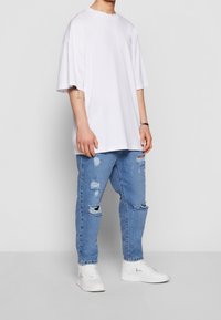 Bílé oversized tričko s krátkými rukávy, kombinované se světle modrými roztrhanými džínami a bílými teniskami. Má volný střih a viditelně ošoupané detaily.