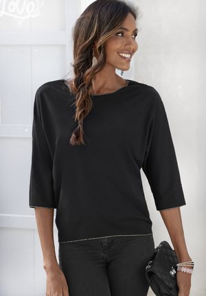 Zwarte, losse top met driekwartmouwen, een ruime halslijn en zilveren stiksel langs de randen. Gepaard met zwarte jeans en een clutch.