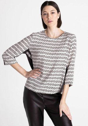 Femme aux cheveux foncés portant un haut blanc orné de cœurs abstraits noirs et un pantalon en cuir noir, posant avec une main sur la hanche.