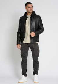 Schwarze Lederjacke mit Kapuze, Reißverschluss und strukturierten Schultern; kombiniert mit dunklen Cargo-Hosen und weißen Sneakers für einen lässigen Look.