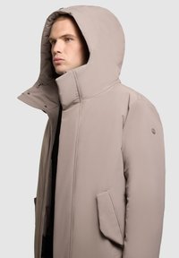 Bézs színű bélelt dzseki nagy kapucnival, magas gallérral és cipzáros záródással. Elöl zsebekkel és sima textúrával rendelkezik. Minimalista kialakítás.