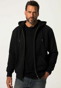 Man met baard, gekleed in een zwart ritsjack en een zwart henley-shirt, staand met één hand in de zak tegen een effen achtergrond.