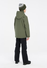 Giacca impermeabile verde con cappuccio, polsini elastici e un branding discreto. Abbinata a pantaloni neri e un berretto a righe verdi e grigie.