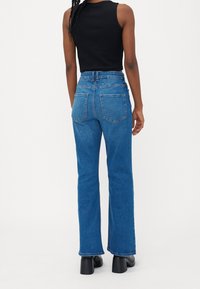 Femme portant un jean bleu taille haute et un haut noir sans manches, debout de dos, sur fond blanc.