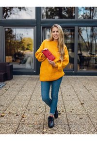 Yellow oversized sweater, blue jeans, and black sneakers; holding a red wallet. Outdoor setting with glass windows in background.