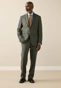 Man with gray beard wearing a green plaid suit, light blue shirt, orange tie, and black dress shoes standing against a beige wall.