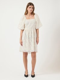 Helles beiges Kleid mit einem strukturierten Blumenmuster, quadratischem Ausschnitt, Puffärmeln und einer gerafften Taille. Kombiniert mit schwarzen spitzen Schuhen.