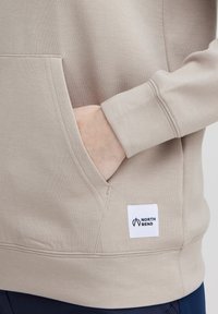 Sweatshirt beige avec une poche frontale et des poignets côtelés. Présente une étiquette blanche avec le logo "NORTH BEND". Le tissu semble doux et extensible.