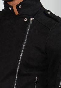 Uomo che indossa una giacca in suede nera con zip diagonale argentata, bottoni a pressione sul colletto e spalline, e tasca con zip sulla manica.