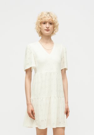 Jeune personne aux cheveux blonds courts et bouclés, portant une robe blanche à manches courtes avec col en V en tissu ajouré et un collier simple, debout, posture neutre.