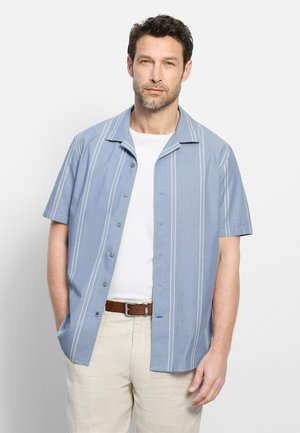 Man die een lichtblauw kort mouw overhemd met witte verticale strepen draagt over een wit T-shirt en beige broek met een bruine riem.