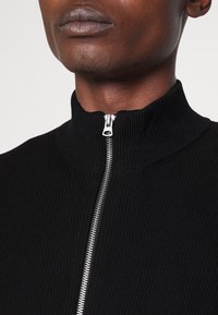 Sudadera negra de cuello alto con relieve y una cremallera plateada en la parte delantera. La tela tiene un aspecto texturizado y un diseño ajustado.