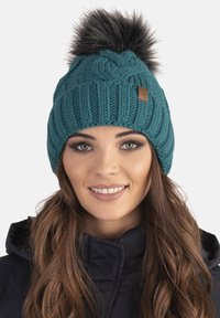 Bonnet en tricot teal avec texture côtelée et un pompon en fausse fourrure sur le dessus. Présente une petite étiquette en cuir de marque sur le côté.