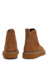 Stivali ankle in suede marrone con linguette, suole di gomma spesse e cuciture visibili, mostrati da dietro su uno sfondo bianco.