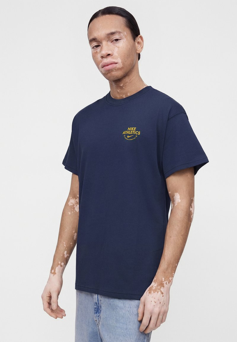 Giovane con vitiligine che indossa una maglietta Nike Athletics blu navy e jeans azzurri, in piedi davanti a uno sfondo chiaro uniforme.