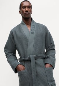 Accappatoio grigio tessuto con colletto a scialle e dettaglio ricamato "BOSS". Presenta una cintura con fiocco e due tasche frontali.