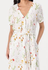 Blommig klänning gjord av lättviktigt tyg, med en vit bakgrund och mönster av gröna och färgglada blommor, korta ärmar och midjebälte.