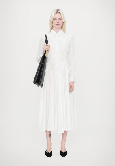 Vestido camisero blanco de manga larga con cierre de botones y cintura fruncida. Los accesorios incluyen un bolso de tote negro texturizado y zapatos de punta negros.