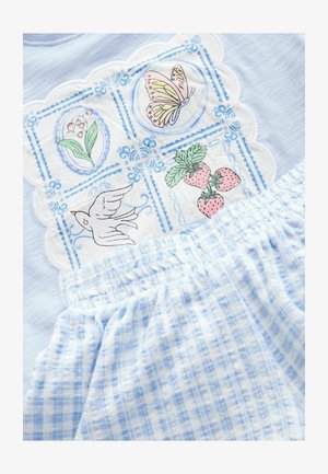 Hellblaue Baumwollbluse mit dekorativen weißen Stickereien, die Schmetterlinge, Blumen und Erdbeermotive zeigen; kombiniert mit karierten blauen Shorts.