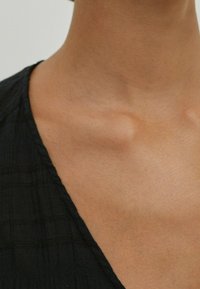 Tessuto nero con un motivo tessurizzato, caratterizzato da un colletto a V, mostrato dalla clavicola fino alla parte superiore del torace. Colore della pelle visibile.