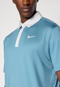 Šviesiai mėlynas polo marškinėliai, pagaminti iš lygios medžiagos, su balta apykakle ir baltu Nike logotipu kairėje krūtinės pusėje. Klasikinio kirpimo dizainas.