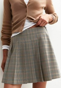 Cárdigan marrón con relieve sobre una blusa blanca, combinado con una falda de cuadros plisada en tonos beiges, negros y marrones. Primer plano de los patrones de la tela.
