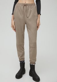 En person bär beigefärgade joggers och svarta ankelskorts, med en figurnära svart långärmad topp, mot en neutral grå bakgrund.