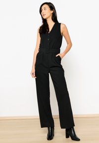 Črna brezrokavnik kombinezon z V-izrezom, sprednjimi gumbi, pasom in širokimi hlačami, v kombinaciji s črnimi čevlji z visokim petam do gležnjev.