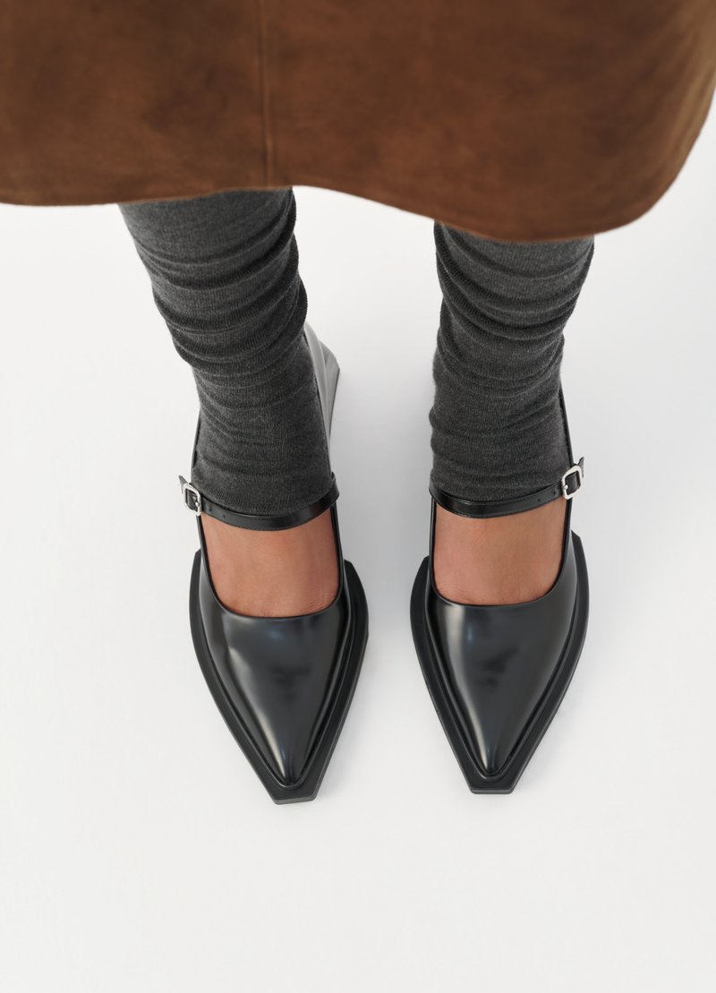 Schwarze Lacklederabsätze mit einer spitzen Zehe, verstellbarem Knöchelriemen und einem niedrigen Blockabsatz, kombiniert mit grauen texturierten Leggings.